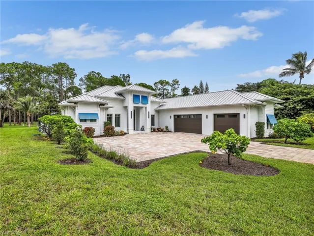 White exterior with blue awnings, a metal roof, a paver driveway, and an attached three-car garage