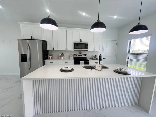 Kitchen with stainless steel appliances, a sink, a center island with sink, light marble finish flooring, and white cabinetry