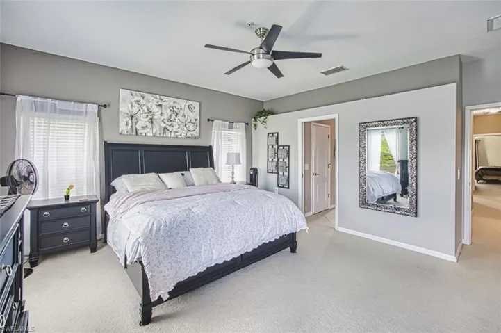 Spacious bedroom featuring light-toned carpeting, neutral wall colors, dual windows with blinds and sheer curtains, a ceiling fan with integrated lighting, and multiple entryways
