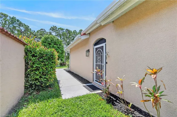 Arched entry door with decorative security screen, stucco exterior, exterior wall lantern, concrete walkway, and established landscaping