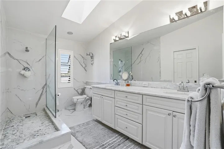 Primary bath featuring a skylight, double vanity, plantation shutters, marble finished shower, and light marble flooring