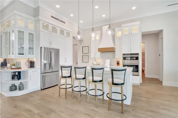 Spacious kitchen featuring an expansive island with seating, pendant lighting, professional-grade stainless steel appliances, abundant white cabinetry with glass-front accents, and wood-finish flooring