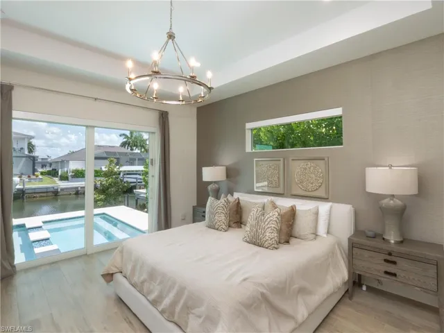Primary Bedroom with Sliding Glass Doors to Pool Area, Light Wood-Finish Flooring, Neutral Textured Wall Finishes, Pendant Chandelier with Integrated Lighting, and Dual Nightstands with Built-In Storage