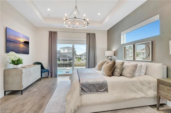 Spacious bedroom featuring wood-finish flooring, a tray ceiling with a contemporary chandelier, and a sliding glass door opening to a waterfront backyard with a pool