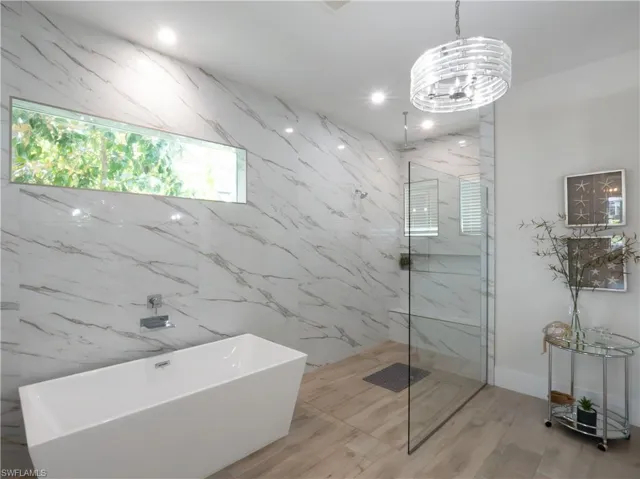 Freestanding soaking tub, walk-in shower with glass enclosure, marble-style wall tiling, wood-finish flooring, and a linear transom window