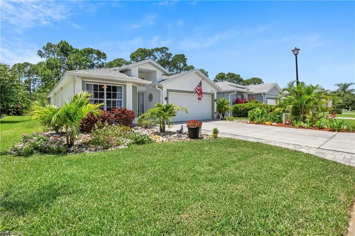 Single-story residence featuring a two-car garage, concrete driveway with paver accents, and a landscaped front yard with mature trees and tropical plantings