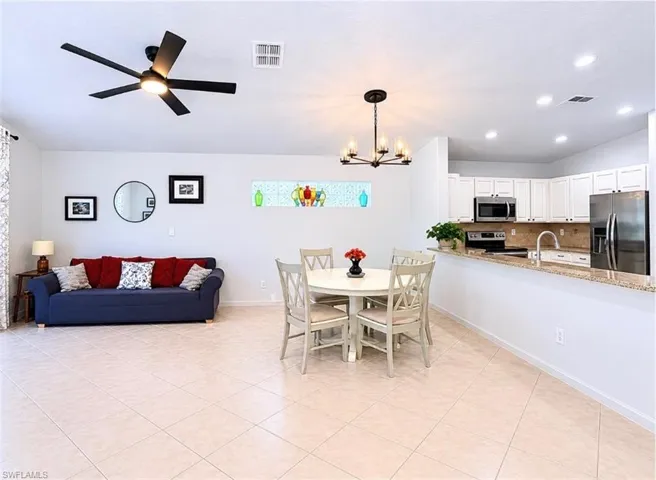 Open-concept living space with tile flooring throughout, featuring a modern ceiling fan, a dining area with a chandelier, and a kitchen with white cabinetry, stainless steel appliances, and a granite-finish countertop with a matching backsplash