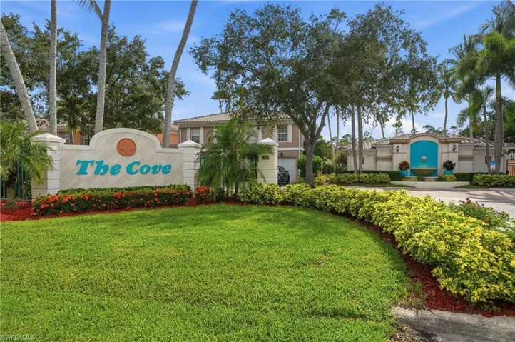 Community entrance featuring a stone sign, lush landscaping, mature palm trees, a decorative fountain, and a stucco-finished wall