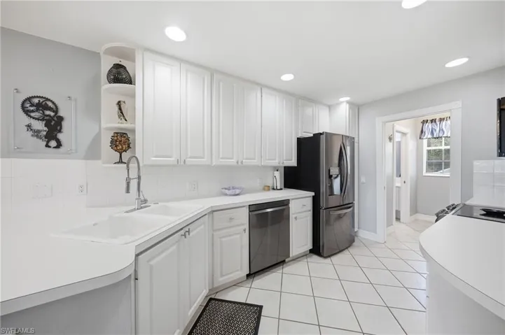 Kitchen featuring white shaker-style cabinetry, a double basin sink with a gooseneck faucet, light-toned countertops, a stainless steel refrigerator, and recessed lighting