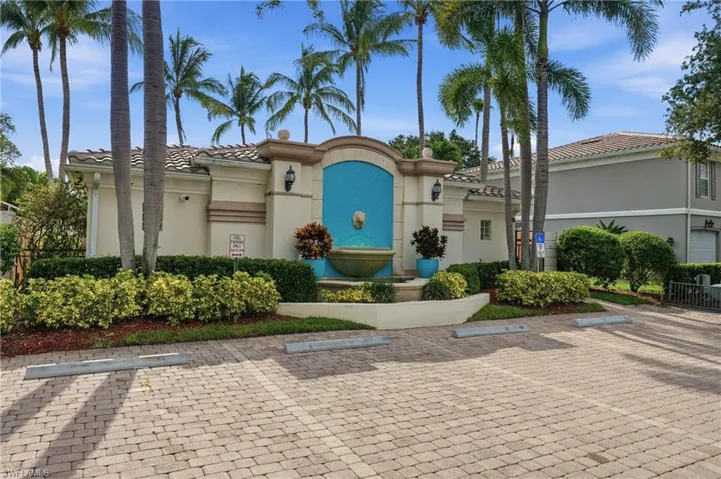Community entrance featuring a stone fountain, lush landscaping, mature palm trees, a decorative fountain, and a stucco-finished wall
