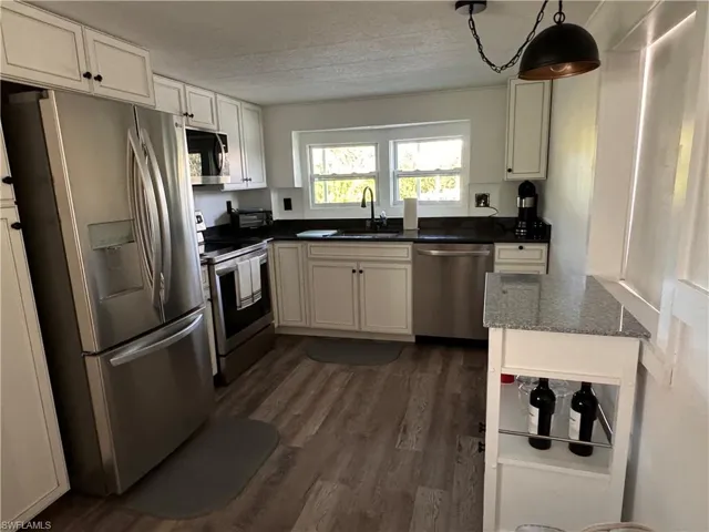 Kitchen with stainless steel appliances, dark wood-style flooring, dark stone counters, white cabinets, and decorative light fixtures