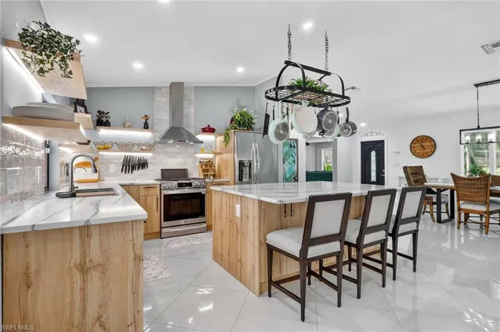 Remodeled kitchen featuring floating shelves, light stone counters, wall chimney exhaust hood, a kitchen island, and appliances with stainless steel finishes