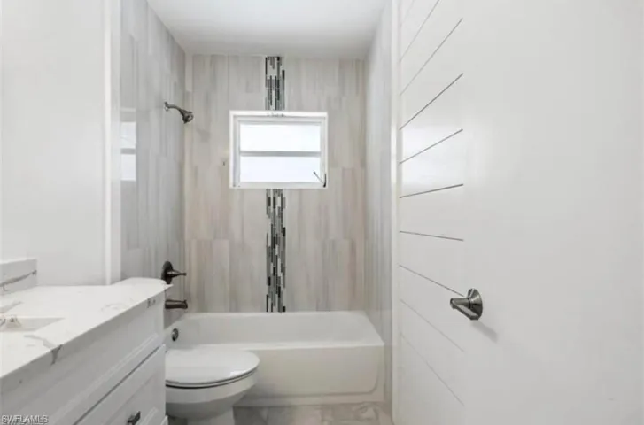 Bathroom featuring a built-in tub with a shower fixture, light-toned vertical wall tiling with a mosaic accent, a small window, a white vanity with a marble-finish countertop, and a toilet