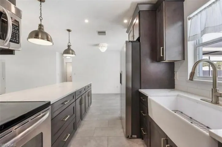 Modern kitchen featuring dark wood cabinetry, stainless steel appliances, a large center island with a light-toned countertop, a farmhouse sink, and contemporary pendant lighting