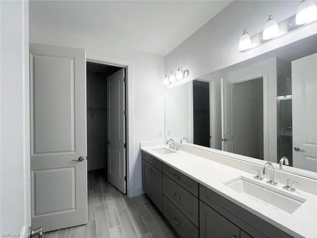Double vanity featuring dark cabinetry, dual rectangular under-mount sinks, light-toned stone-finish countertops, full-width mirror, and multi-bulb light fixtures