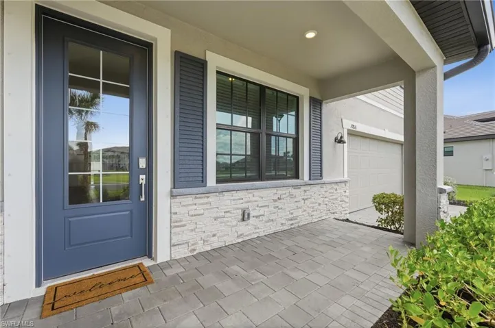 Covered front entry featuring paver flooring, a paneled front door with glass inserts, stone accent siding, and exterior wall sconces