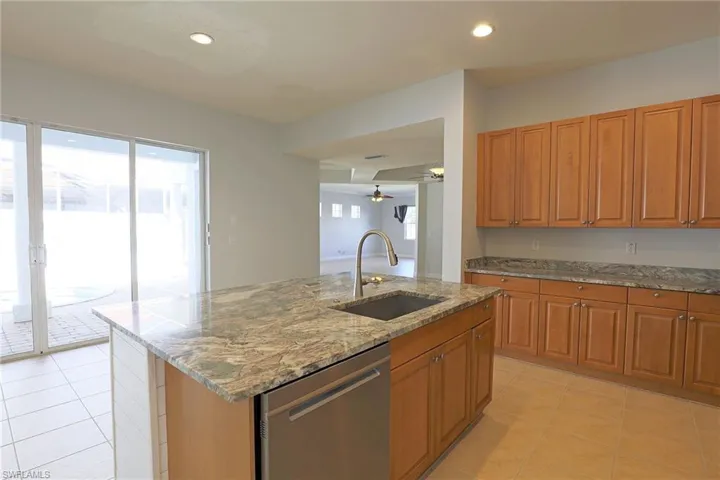 Kitchen featuring wood cabinetry, stone countertops, central island with undermount sink, stainless steel dishwasher, and ceramic tile flooring