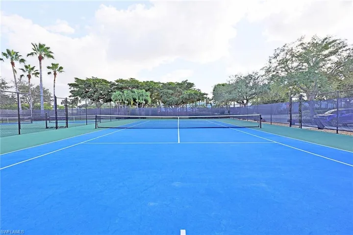 Blue-surfaced tennis court featuring a perimeter chain-link fence and mature palm trees