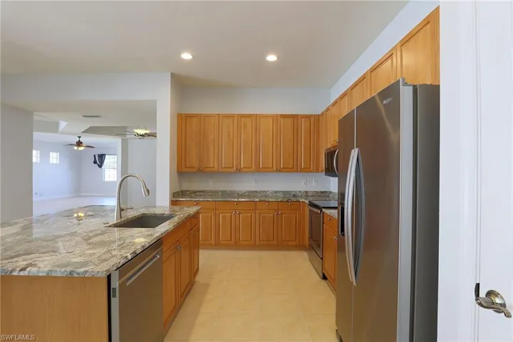 Kitchen featuring wood cabinetry, stone countertops, an island with an integrated stainless steel sink, tile flooring, and stainless steel appliances