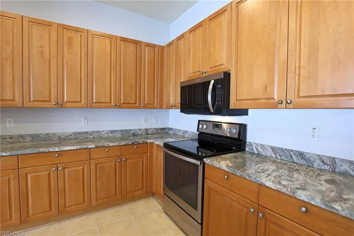 Kitchen featuring wood cabinetry, natural stone countertops, tile flooring, and stainless steel appliances including an over-the-range microwave and electric range