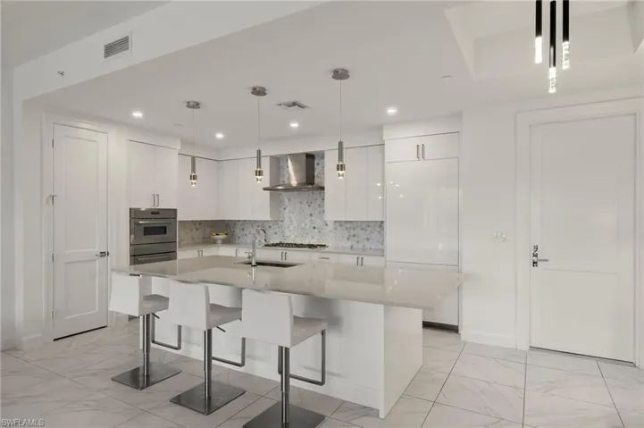 Kitchen featuring white cabinetry, an island with sink, wall chimney range hood, and decorative light fixtures