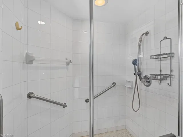 Shower enclosure with white tiled walls, glass paneling, a hand-held shower fixture, and grab bars