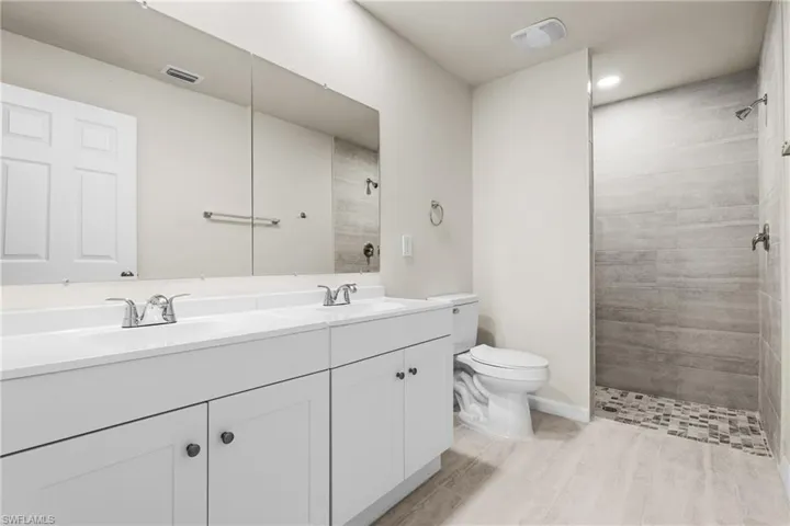 Bathroom featuring a double vanity with white cabinetry, a large wall-mounted mirror, a walk-in shower with textured tile, a mosaic shower floor, and light-toned floor tile
