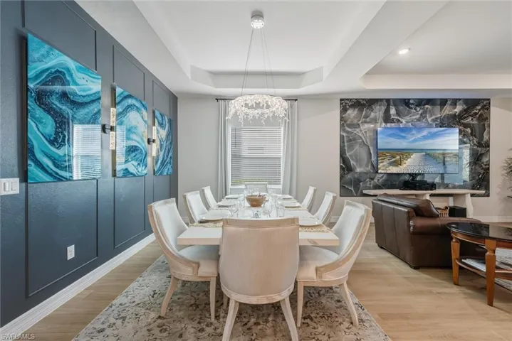 Luxurious Dining area with an ultra-modern chandelier, decorative custom wall, a tray ceiling, large window with blind and modern drapes, and light wood-style plank tile flooring