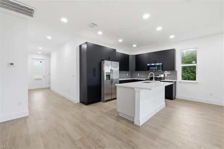 Modern kitchen featuring dark cabinetry, stainless steel appliances, a white island with a sink, grey subway tile backsplash, and light wood-finish flooring