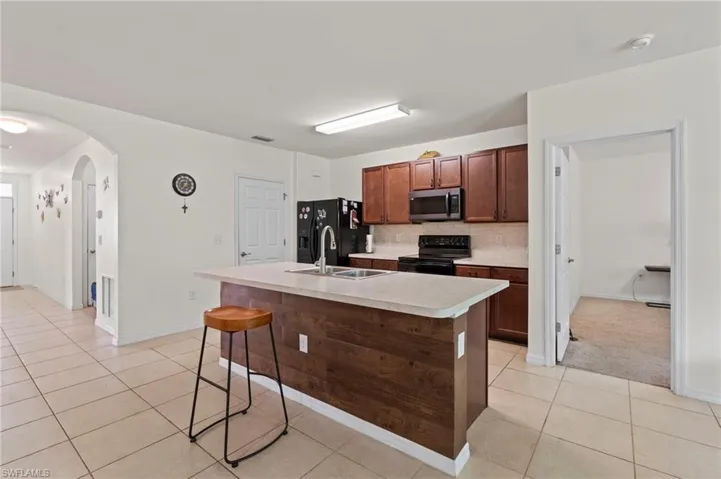 Kitchen featuring a center island with sink, light tile patterned floors, black appliances, light countertops, and arched walkways