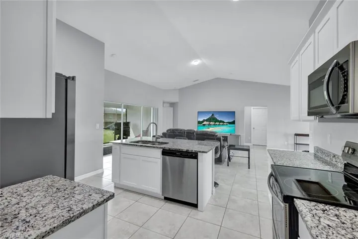 Kitchen featuring appliances with stainless steel finishes, white cabinets, lofted ceiling, a center island with sink, and light stone counters