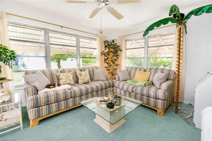 Carpeted living area featuring multiple windows with horizontal blinds, a ceiling fan, and a glass-top coffee table with a stone-finish base