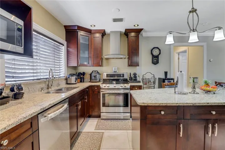 Kitchen with light stone counters, wall chimney exhaust hood, stainless steel appliances, pendant lighting, and a sink