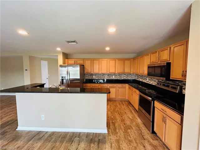 Kitchen featuring stainless steel appliances, an island with sink, light wood-style flooring, backsplash, and dark stone countertops