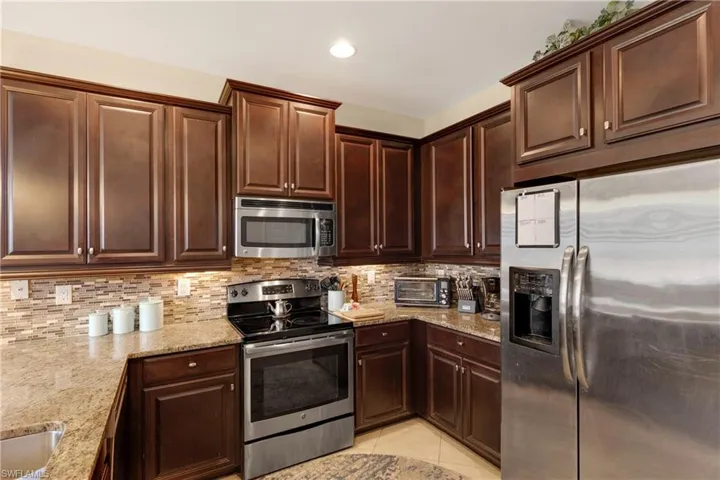 Kitchen with light tile patterned floors, dark brown cabinets, stainless steel appliances, light stone counters, and tasteful backsplash
