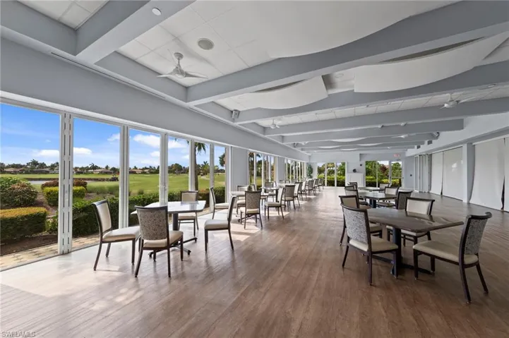 Expansive open-concept dining and lounge area featuring floor-to-ceiling glass doors and windows showcasing pastoral grounds, wide-plank wood-finish flooring throughout, coffered ceiling with recessed lighting, and contemporary upholstered seating arrangements positioned to maximize natural light and sightlines