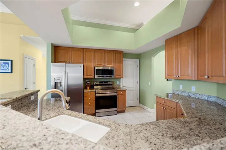 Kitchen featuring stainless steel appliances, a raised ceiling, light stone counters, wood finish cabinetry, and arched walkways