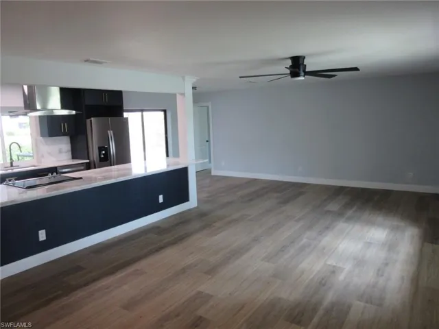 Kitchen with stainless steel refrigerator with ice dispenser, a ceiling fan, wall chimney range hood, open floor plan, and dark cabinets