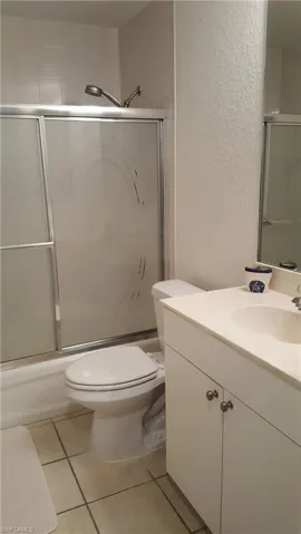Bathroom with vanity, a textured wall, toilet, bath / shower combo with glass door, and tile patterned flooring