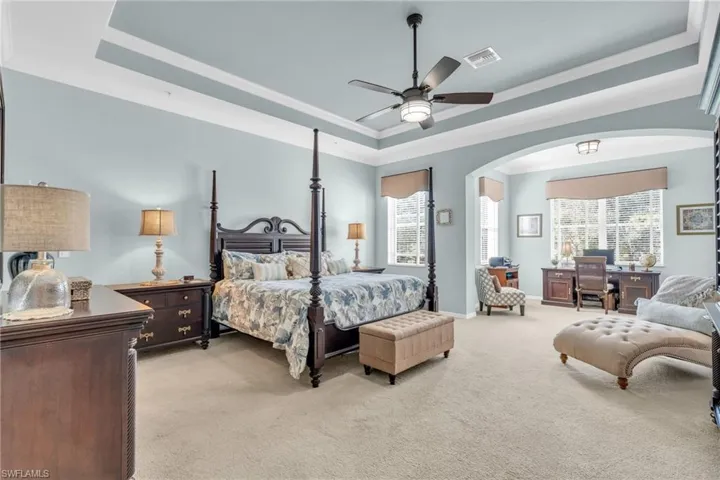 Bedroom featuring a raised ceiling, light carpet, ornamental molding, multiple windows, and ceiling fan
