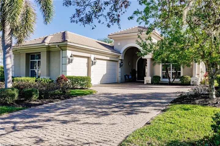 Stucco exterior with a tile roof, paver driveway, arched entryway, and mature landscaping