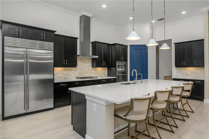 Kitchen with a kitchen breakfast bar, wall chimney exhaust hood, hanging light fixtures, a center island with sink, and stainless steel appliances