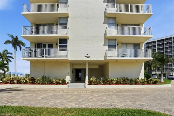 Multi-story residential building featuring private balconies with white railings, a paver driveway, and ground-level landscaping - Virtually Edited Image