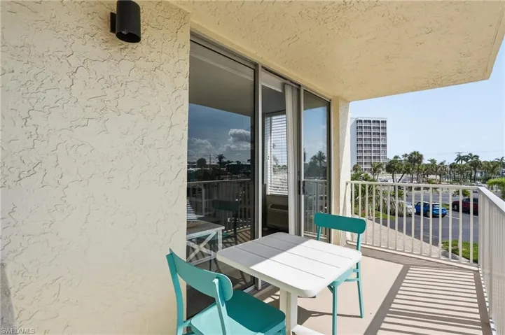 Private balcony featuring textured stucco walls, sliding glass doors with vertical blinds, a white metal railing, and a partial cityscape with palm trees - Virtually Edited Image