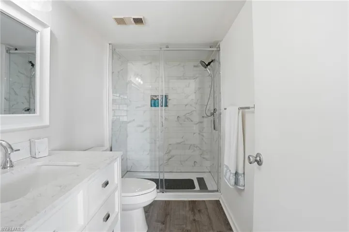 Bathroom featuring a glass-enclosed shower with subway tile surround, a single vanity with an undermount sink and light-toned countertop, wood-finish flooring, and a framed mirror - Virtually Edited Image