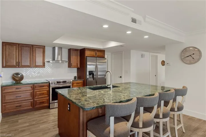 Kitchen featuring wall chimney exhaust hood, sink, an island with sink, stainless steel appliances, and decorative backsplash