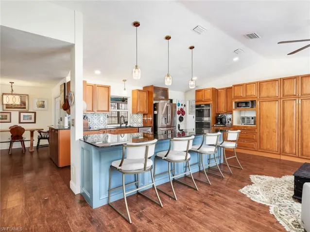 Spacious kitchen featuring a large island with a blue base and seating, dark stone countertops, wood-finish flooring, ample wood cabinetry, and a patterned tile backsplash
