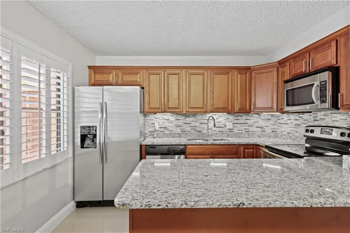 Kitchen with light stone countertops, appliances with stainless steel finishes, a textured ceiling, light tile patterned flooring, and brown cabinetry