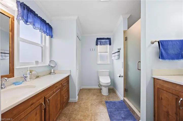Dual-sink wooden vanity with light-toned solid surface countertops, tile flooring, glass-enclosed shower, and two windows