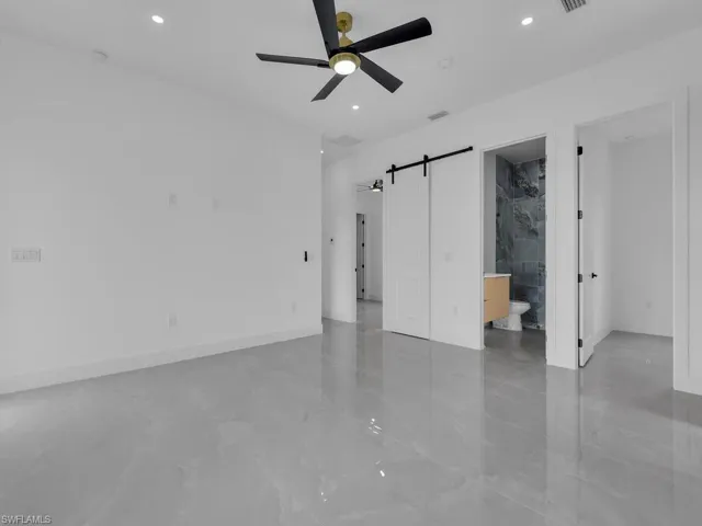 Unfurnished bedroom with a barn door, ceiling fan, ensuite bathroom, recessed lighting, and light marble finish flooring